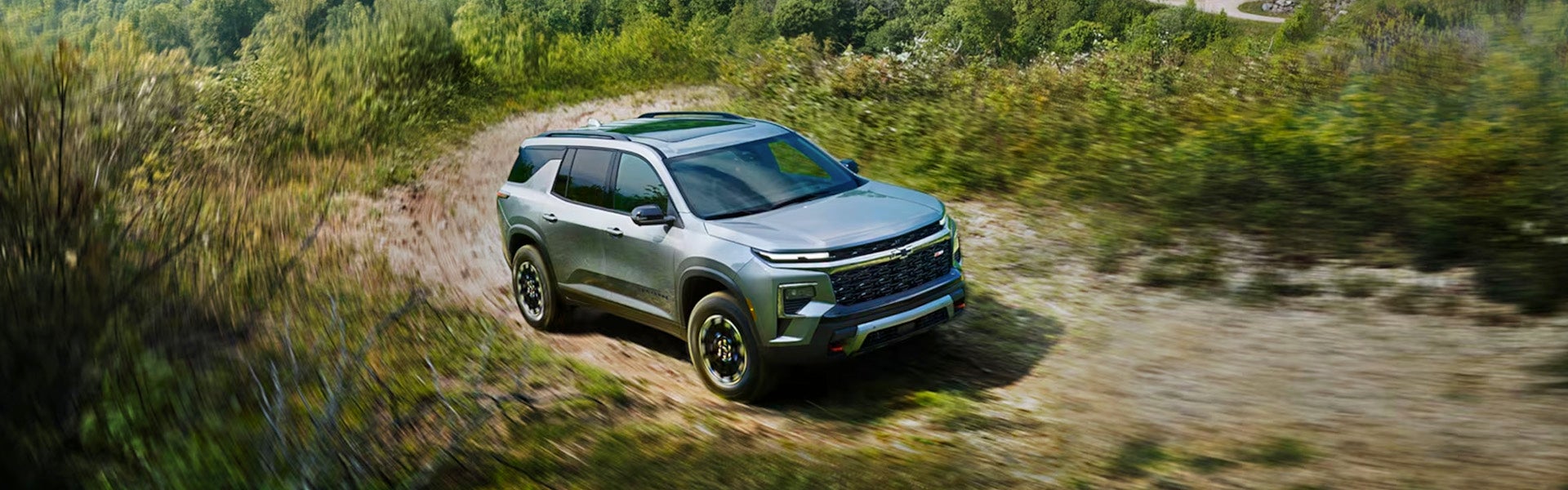 A silver 2024 Chevrolet Traverse driving on an off-road dirt path with greenery in the background