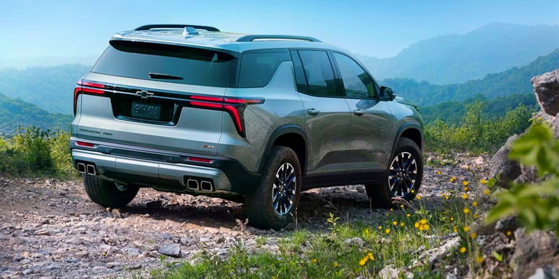 A rear-side view of the 2024 Chevrolet Traverse driving on a rugged trail with mountains in the background
