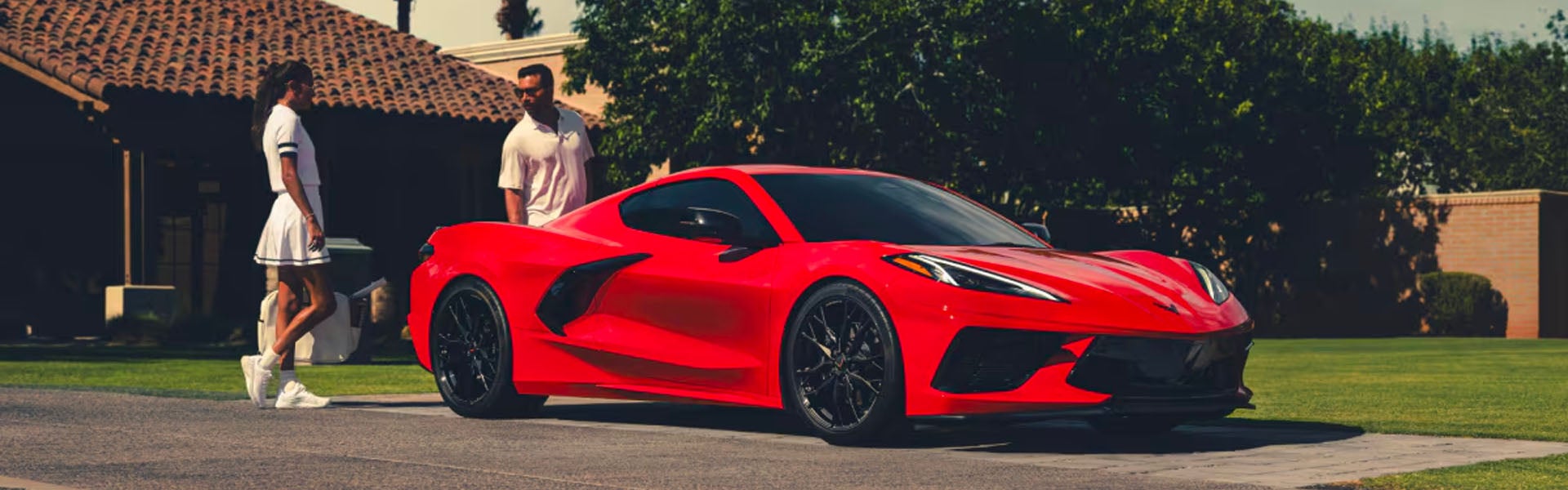 A red 2025 Chevrolet Corvette Stingray parked in front of a modern house with three people nearby